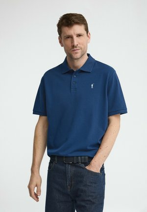 Man die een marineblauw poloshirt met korte mouwen en een klein wit logo draagt, donkere jeans en een zwarte riem, staand met één hand in de zak.