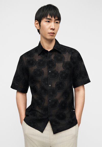 Μαύρο διαφανές πουκάμισο με κοντά μανίκια, με υφή και λουλουδάτο σχέδιο, μπροστινά κουμπιά και γιακά, συνδυασμένο με ανοιχτόχρωμο παντελόνι.