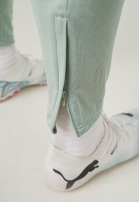 Mintgroene sportbroek met een textuurpatroon, voorzien van een zijrits bij de enkel. Witte sportschoenen met een zichtbaar zwart logo.