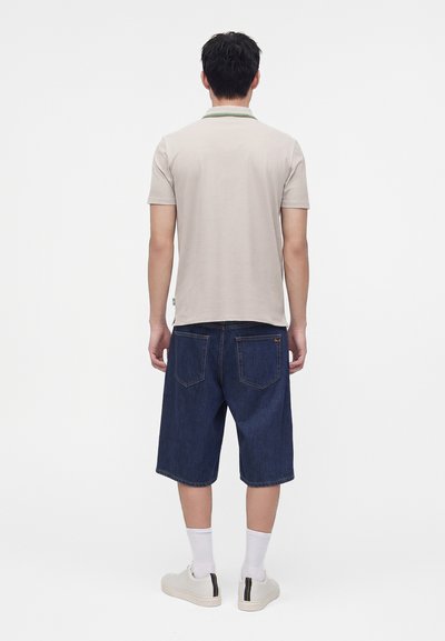 Homme portant un polo beige à manches courtes, un short en jean bleu foncé jusqu'aux genoux, des chaussettes blanches et des baskets blanches, debout de dos.
