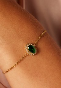 Bracelet en chaîne dorée avec une gemme verte ovale entourée de pierres claires circulaires, présenté sur un poignet bronzé.