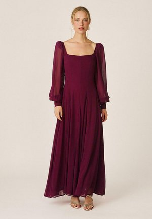 Mujer con un vestido largo color ciruela con mangas abullonadas transparentes y escote cuadrado, de pie con las manos relajadas a los lados.