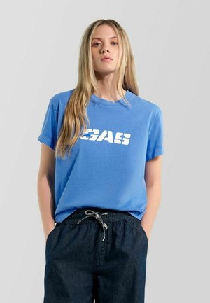 Junge Frau mit langem blondem Haar, die ein blaues T-Shirt mit weißem Schriftzug und dunkle Hosen mit Kordelzug trägt, steht mit den Händen in den Taschen.