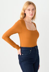 Body z długim rękawem i kwadratowym dekoltem w kolorze rdzawego pomarańczu, wykonany z gładkiej, elastycznej tkaniny. W zestawieniu z jeansami o wysokim stanie w ciemnym odcieniu niebieskiego.