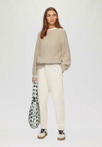Beige gebreide trui met lange mouwen, witte shirt eronder, crèmekleurige sweatpants, zwart-wit gestreepte tas en beige sneakers.