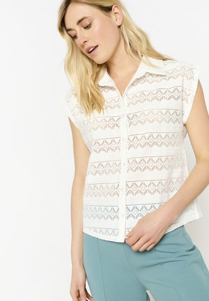Chemise blanche à boutons avec un tissu en dentelle et des découpes géométriques, manches courtes et col pointu. Portée avec un pantalon bleu clair.