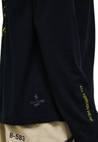 Schwarzes Sportshirt aus strukturiertem Material, mit Sorona®-Logo und "ALL TERRAIN WEAR" in Gelb auf dem Ärmel.