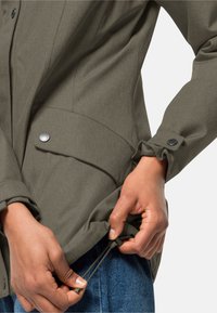 Olivgrön jacka med en strukturerad finish; har knappar, sidofickor och en dragsko i nederkant som justeras med händerna.