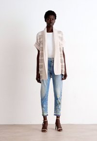 Strickjacke in hellbeige mit offenem Vorderteil, kurzen Ärmeln und strukturierten Streifen. Kombiniert mit hellblauen Jeans mit floralen Stickereien.
