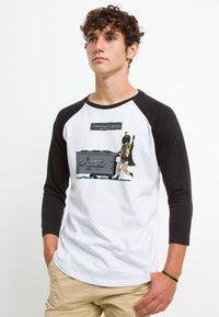 T-shirt raglan bianco e nero con un'illustrazione grafica di un personaggio che trascina una valigia grigia con il testo "Bagaglio sovradimensionato."