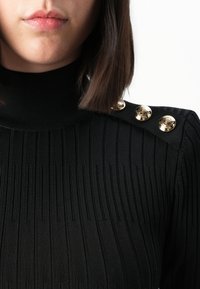 Donna che indossa un maglione a collo alto nero a coste con tre bottoni decorativi dorati sulla spalla, visibili dal mento al petto.