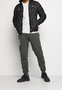 La chaqueta de plumas negra cuenta con capucha y un patrón de acolchado diagonal, combinada con pantalones jogging de color verde oliva. Las zapatillas negras completan el atuendo.