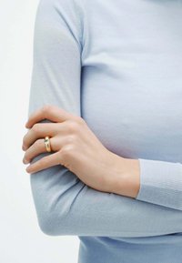 Anillo de oro con una piedra central clara, llevado en una mano, sobre un suéter azul claro de textura suave y diseño ajustado.