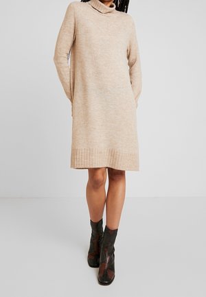 Kobieta w beżowej, długorękawowej sukience z dzianiny z golfem, w połączeniu z czarno-brązowymi botkami w wzory, stojąca na jednolitym tle.