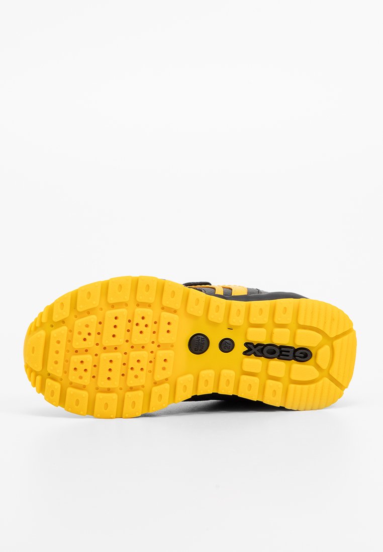 Semelle de chaussure de sport noire avec des motifs de trame profonds, présentant une semelle extérieure jaune vif et un logo "GEOX" noir. Caoutchouc texturé pour une meilleure traction.