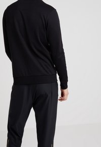 Svart långärmad sweatshirt med rund halsringning, mjukt material och ribbad fåll, bärs med svarta byxor med en subtil sidostrimma.