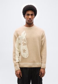 Άνδρας με αφροκρέμασμα που φοράει μπεζ πλεκτό πουλόβερ με ανάγλυφη λευκή σχεδίαση κεφαλιού αλόγου, συνδυασμένο με μαύρο παντελόνι σε απλό φόντο.