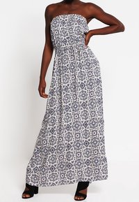 Kvinna i ärmlös, axelbandslös vit maxiklänning med svarta geometriska mönster och svarta öppna klackskor, poserar med handen på höften.