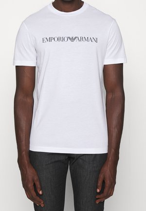 Homme portant un t-shirt blanc à manches courtes avec le logo "EMPORIO ARMANI" en noir centré sur la poitrine, associé à un pantalon foncé.