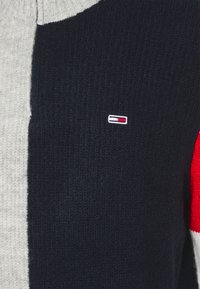 Stickad tröja med hög krage, med grå, marinblå och röda färgblock. Liten logotyp i mitten, ribbad textur genom hela tröjan.