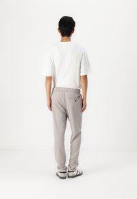 Graue Sweatpants mit elastischem Bund, einer Gesäßtasche und einem weißen T-Shirt mit kurzen Ärmeln werden getragen. Die Schuhe sind schwarz und weiß mit einer strukturierten Sohle.