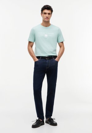 Mees seisab käed taskutes, kannab helesinist Tommy Hilfigeri T-särki, tumedatesiniseid teksapükse, musta vööd ja musti kingi valgel taustal.