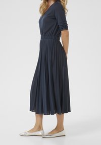 Robe bleu marine plissée avec un décolleté en V et des manches trois-quarts, associée à des ballerines en argent. Tissu lisse avec une taille élastique.