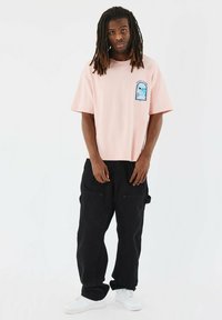 Camiseta rosa de manga corta con un parche gráfico en el pecho, combinada con pantalones cargo negros y zapatillas blancas. Ajuste casual y tejido.