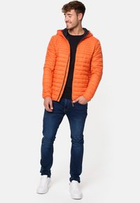 Oranssi tikattu takki hupulla, jossa on kontrastina tumma sisäosa, vetoketjukiinnitys, käytössä mustan paidan ja sinisten farkkujen päällä, valkoiset tennarit.
