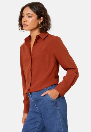 Rostfarbige Bluse mit langen Ärmeln, Kragen, Fronttasche und knöpfbaren Manschetten, kombiniert mit hochtaillierten blauen Jeans. Glatte Stofftextur.