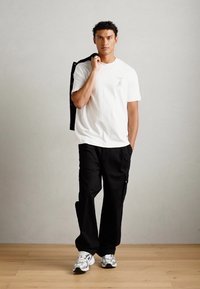 Witte T-shirt met grafische details, zwarte losse broek en witte sneakers. Een extra zwarte jas is over de schouder gedrapeerd.