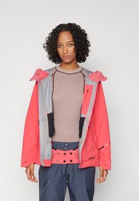 Rote und graue wasserdichte Jacke mit Kapuze, ausgestattet mit Reißverschlusstaschen und einem strukturierten braunen Strickpullover darunter. Marineblaue Hose rundet das Outfit ab.
