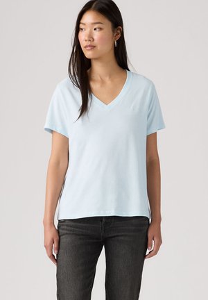 Kvinde med langt mørkt hår iført en lyseblå V-hals kortærmet skjorte og sorte jeans, kiggende mod venstre mod en ensfarvet baggrund.