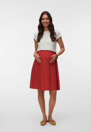Mujer embarazada sonriente con cabello largo y oscuro, lleva una blusa blanca, falda roja y zapatos planos marrones, de pie con las manos en su vientre.