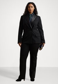 Čierny strihaný dvojradový blejzer so saténovými klopami, doplnený o zladené čierne nohavice. Modelka drží modrú a zlatú kabelku.
