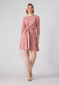 Knelang rosa kjole med trekvart ermer, med en knyttet midje detalj og en teksturert stoff. Modellen har på seg beige stroppeskinn.