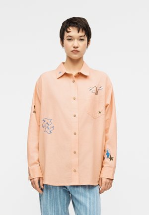 Νεαρό άτομο με κοντά σκούρα μαλλιά, που φοράει ένα άνετο ροδακινί πουκάμισο με κεντημένα πουλιά, λουλούδια και αστέρια, σε συνδυασμό με μπλε ριγέ παντελόνια.