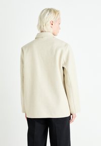 Beige ullblandingsfrakk med strukturelt design, rund krage og rett snitt. Har synlige sømmer og knappelukking bak.