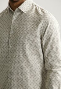 Lange mouwen shirt van een lichtbeige stof met een geometrisch patroon in donkere tinten, met een knoopsluiting bij de kraag en een subtiele textuur op het oppervlak.