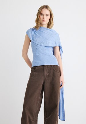 Žena s vlnitými blond vlasmi nosí svetlomodrý vrch s bodkami na jedno rameno s dlhou visiacu látkou a voľné hnedé nohavice.