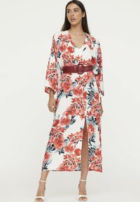 Maxikleid aus weißem Stoff mit rot-blauem Blumenmuster, Kimonoärmeln, V-Ausschnitt und einem hohen Schlitz bis zum Oberschenkel, tailliert mit einem gewebten roten Gürtel.