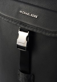 Črn usnjen nahrbtnik z teksturiranim finišom, srebrno kovinsko zaponko in blagovno znamko z napisom "MICHAEL KORS" na pokrovu.