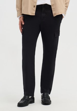 Uomo che indossa pantaloni cargo neri a gamba dritta con tasche laterali, mocassini neri, calzini bianchi, giacca beige e camicia bianca.