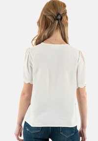 Blouse blanche à texture avec des manches courtes et bouffantes et un encolure arrondie. Associée à un jean bleu foncé et des cheveux longs maintenus par une pince.
