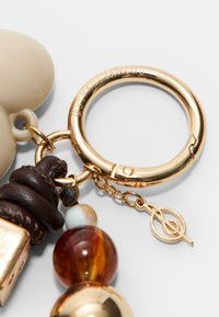Porte-clés couleur or avec un charm beige, des accents en cuir marron et des perles multicolores, ainsi qu'un logo décoratif sur l'anneau.