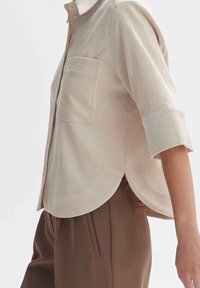 Beige Cord-Hemd mit aufgerollten 3/4-Ärmeln und Fronttasche, kombiniert mit hochgeschnittenen, braunen, plissierten Hosen.