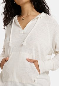 Haut à capuche blanc léger avec un col en V, des cordons de serrage, une poche avant et des poignets côtelés. Tissu texturé avec des variations subtiles de teinte.