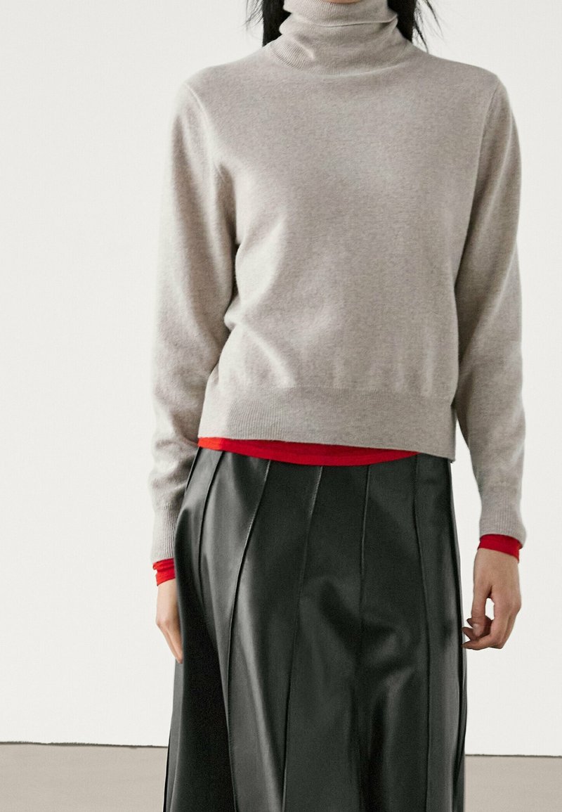 Hellgrauer Rollkragenpullover aus weichem Stoff, tailliert geschnitten. Kombiniert mit einem schwarzen plissierten Rock aus Kunstleder und einem roten Langarmshirt darunter.