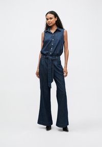 Denim jumpsuit with sleeveless design, button front, belt at the waist, and wide-leg pants. Fabric is a medium-dark blue with smooth texture.