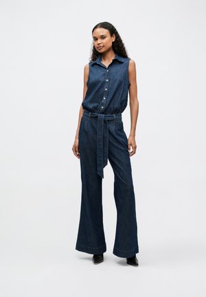 Macacão de ganga com design sem mangas, frente com botões, cinto na cintura e calças largas. O tecido é de um azul médio-escuro com uma textura suave.